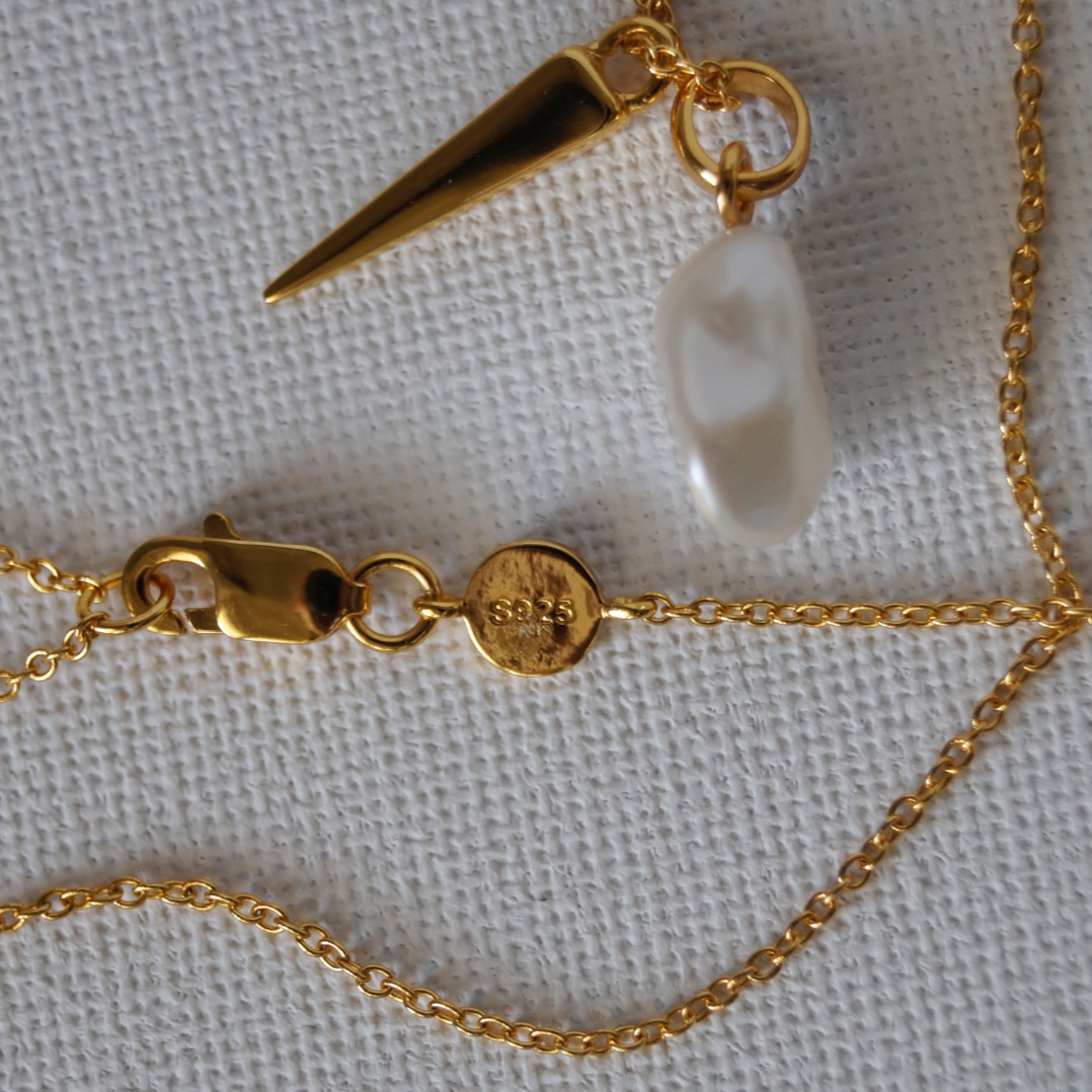 Potvrda autenticnosti.
Zlatna ogrlica s tankim lančićem i dvama privjescima – prirodnim baroknim biserom nepravilnog oblika i zlatnim izduženim detaljem.