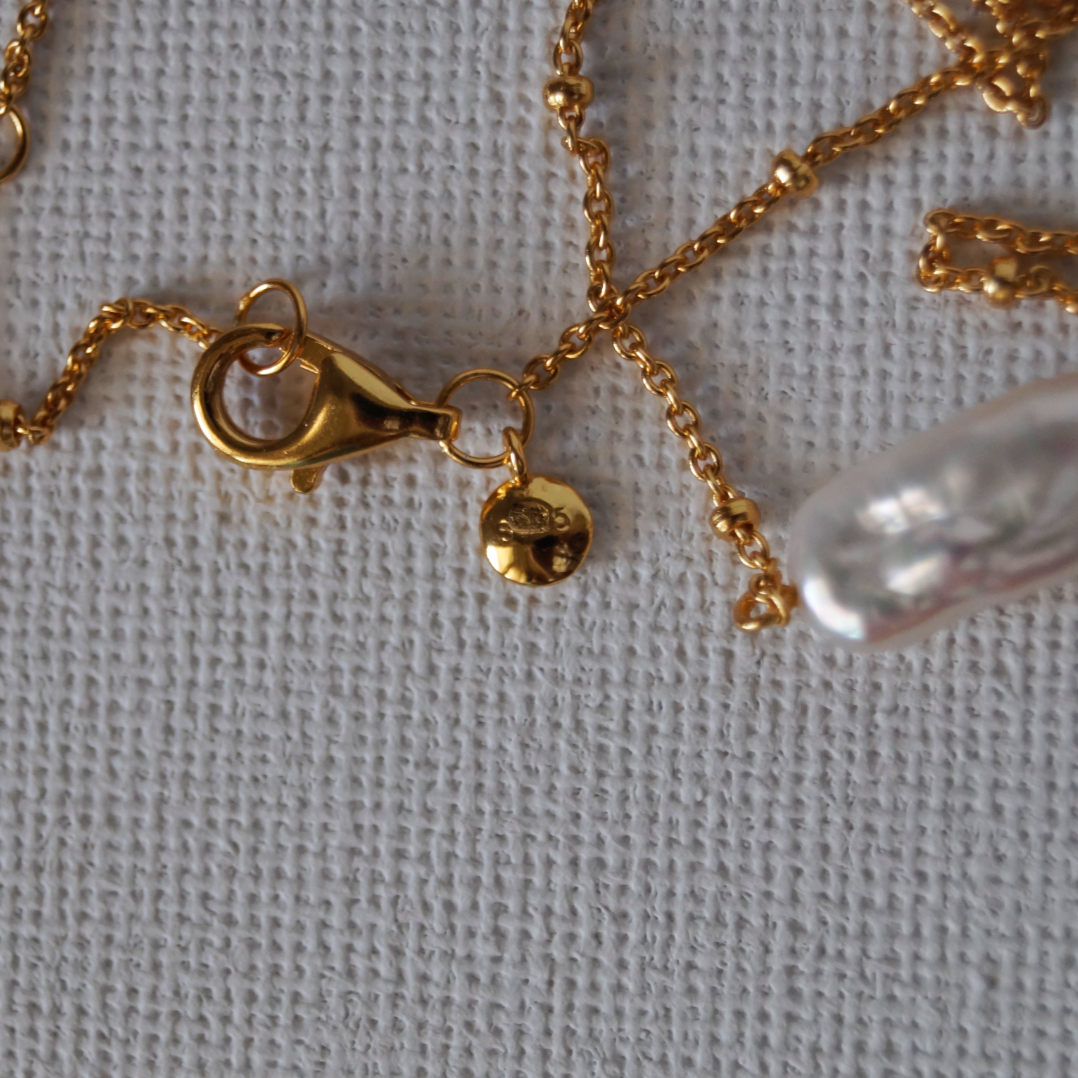 Potvrda autenticnosti.
Zlatna ogrlica sa suptilnim kugličastim lančićem i privjeskom od nepravilnog, svjetlucavog keshi bisera zakrivljenog oblika koji podsjeća na polumjesec.