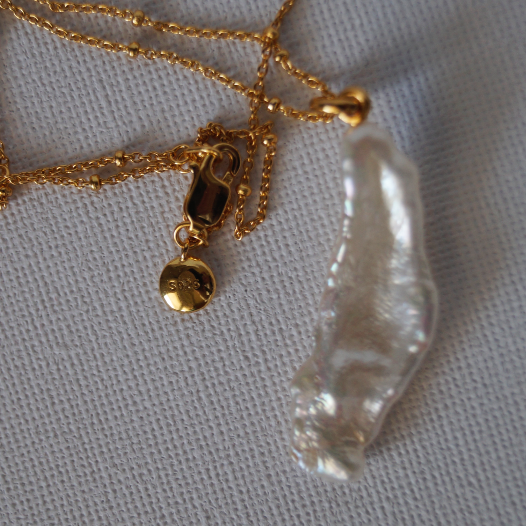 Zlatna ogrlica s tankim lančićem ukrašenim diskretnim kuglicama i nepravilnim keshi biserom visokog sjaja, prikazana na neutralnoj podlozi.