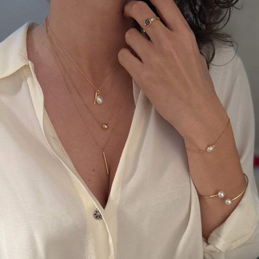 Zlatna ogrlica s tankim lančićem i dvama privjescima – prirodnim baroknim biserom nepravilnog oblika i zlatnim izduženim detaljem.